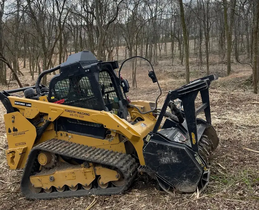 Detailed close-up of a rugged forestry mulching head attached to an excavator's hydraulic arm, resting on fresh wood chips with sharp carbide teeth visible.