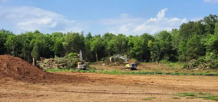 Wide view of a large-scale land clearing operation featuring two Volvo excavators, a massive dirt mound, and cleared property against a lush green forest line.
